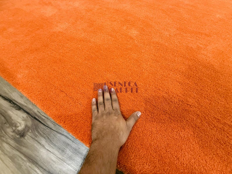 May include: A solid bright orange area rug with a dense, plush, and soft-textured pile. The surface features a small, dark, rectangular logo stamp in the center that reads SENECA CARPET in a clear, sans-serif font, indicating the brand name.