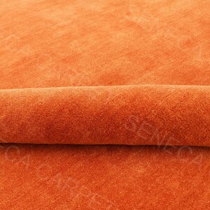 May include: A solid orange carpet with a soft, dense pile texture. The carpet is partially rolled over itself, creating a horizontal fold across the center. The text SENECA CARPET is visible as a watermark repeated across the surface of the fabric.