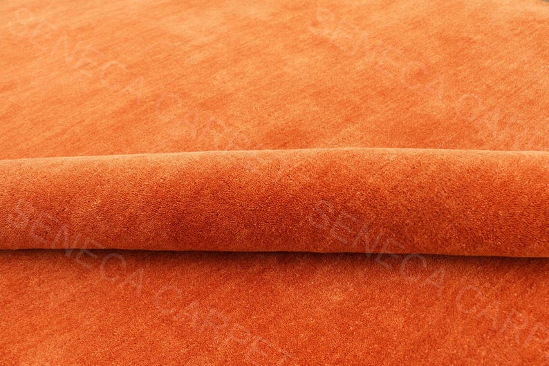 May include: A solid orange carpet with a soft, dense pile texture. The carpet is partially rolled over itself, creating a horizontal fold across the center. The text SENECA CARPET is visible as a watermark repeated across the surface of the fabric.