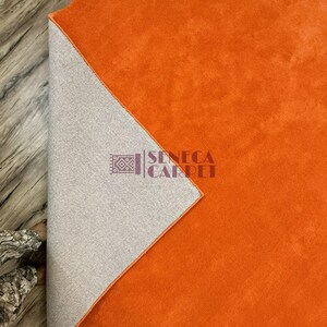 May include: A vibrant orange area rug with a soft, solid-colored surface. One corner is folded back to reveal a light gray, textured backing material. The logo for Seneca Carpet is printed in dark purple text on the gray backing section.