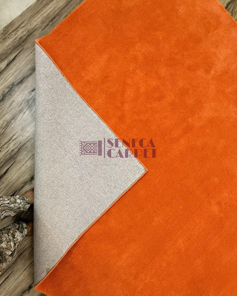 May include: A vibrant orange area rug with a soft, solid-colored surface. One corner is folded back to reveal a light gray, textured backing material. The logo for Seneca Carpet is printed in dark purple text on the gray backing section.