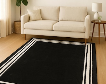 May include: A rectangular black rug with a white border and two white stripes. The rug is placed on a light wood floor in a living room setting, with a beige sofa and a potted plant in the background.