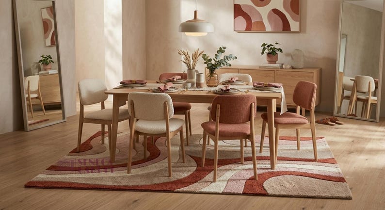 May include: A rectangular area rug featuring a wavy, abstract pattern in shades of terracotta, cream, and soft pink. The rug is placed under a light wood dining table with six chairs, alternating between cream and terracotta upholstery colors.