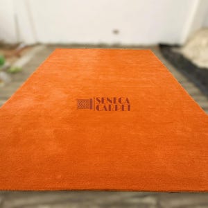 May include: A rectangular, solid orange area rug with a soft, plush texture. The center of the rug features a dark brown logo with a geometric icon and the text SENECA CARPET printed in a clear, sans-serif font across the surface.