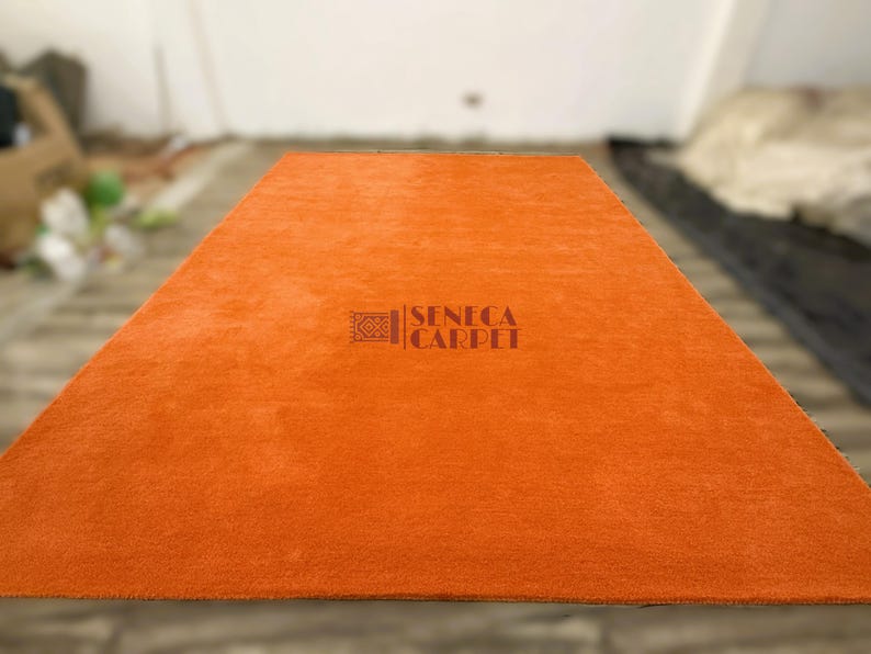 May include: A rectangular, solid orange area rug with a soft, plush texture. The center of the rug features a dark brown logo with a geometric icon and the text SENECA CARPET printed in a clear, sans-serif font across the surface.