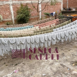 Rows of white, pink, and green yarn hang drying outdoors on lines, with a brick wall and trees in the blurred background.
