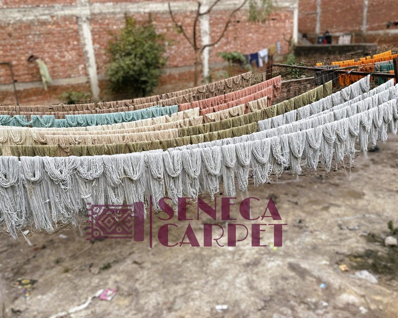 Rows of white, pink, and green yarn hang drying outdoors on lines, with a brick wall and trees in the blurred background.
