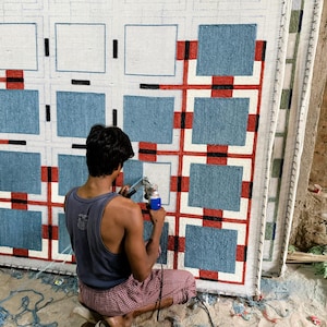 May include: A large rectangular rug featuring a grid pattern of blue squares separated by white borders, accented with red and black lines. The rug is mounted on a frame, and a tufting gun is being used to fill in the white sections with yarn.