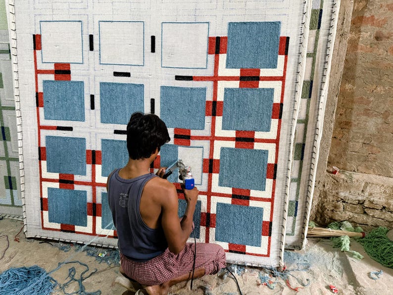 May include: A large rectangular rug featuring a grid pattern of blue squares separated by white borders, accented with red and black lines. The rug is mounted on a frame, and a tufting gun is being used to fill in the white sections with yarn.