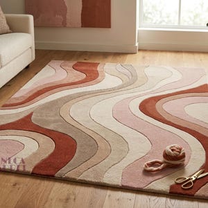May include: A rectangular area rug featuring a wavy, multi-colored pattern in shades of terracotta, dusty rose, beige, and cream. The rug has a textured, carved appearance with flowing lines and includes the text SENECA CALDEL in the bottom corner.