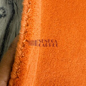 May include: A vibrant orange carpet with a thick, plush pile texture. The edge of the carpet shows a dense, bound finish. The text SENECA CARPET is printed in a dark color on the surface of the orange fabric, accompanied by a small geometric logo.