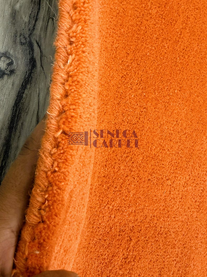 May include: A vibrant orange carpet with a thick, plush pile texture. The edge of the carpet shows a dense, bound finish. The text SENECA CARPET is printed in a dark color on the surface of the orange fabric, accompanied by a small geometric logo.