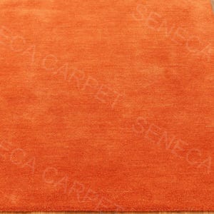 May include: A solid orange area rug with a dense, short-pile texture. The surface features a repeating watermark text overlay that reads SENECA CARPET in a light, semi-transparent font across the entire fabric of the rectangular floor covering.