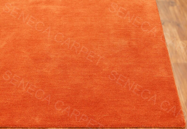 May include: A solid orange area rug with a dense, short-pile texture. The surface features a repeating watermark text overlay that reads SENECA CARPET in a light, semi-transparent font across the entire fabric of the rectangular floor covering.
