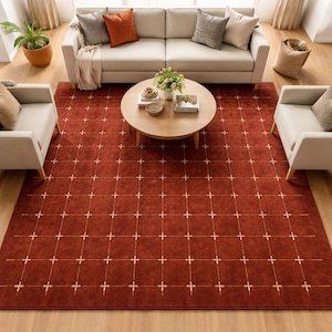 Pode incluir: Um tapete vermelho com um padrão de grade de cruzes brancas. O tapete está em uma sala de estar com um sofá bege, duas poltronas e uma mesa de centro redonda de madeira. Almofadas decorativas e uma cesta também são visíveis.