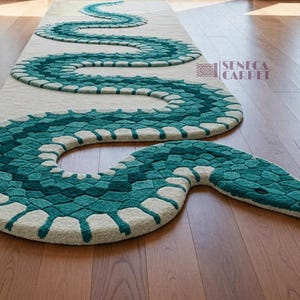 Op de afbeelding: Een teal en wit slangvormig tapijt met een textuurontwerp. Het tapijt ligt op een hardhouten vloer en heeft een lange, kronkelende vorm. De kop van de slang bevindt zich rechtsonder, met een blauwe tong.