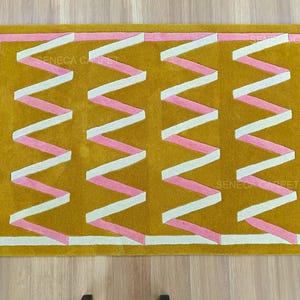 May include: Mustard yellow rectangular rug with a geometric design. The rug features a pink and white zigzag pattern across the center, framed by a pink and white border. The text "SENECA CARPET" is visible on the rug.