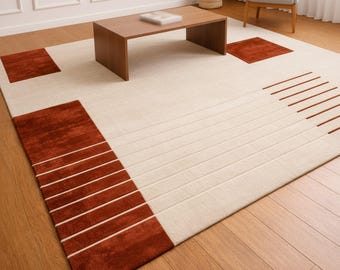 Puede incluir: Una alfombra rectangular grande con una base de color crema y acentos geométricos en un tono naranja óxido profundo. El diseño incluye bloques cuadrados sólidos en las esquinas y líneas horizontales paralelas que crean un patrón de rayas.
