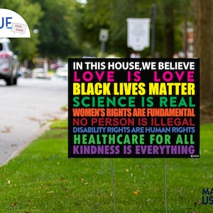 May include: Black yard sign with white text that reads "In this house, we believe: Love is love, Black Lives Matter, Science is real, Women's rights are fundamental, No person is illegal, Disability rights are human rights, Healthcare for all, Kindness is everything."