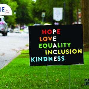 May include: Black yard sign with the words "HOPE", "LOVE", "EQUALITY", "INCLUSION", and "KINDNESS" in rainbow colors. The sign is on a green lawn with a car in the background. The sign also says "MADE IN USA" at the bottom.