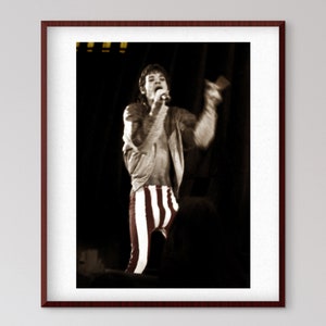 May include: A black and white photograph of a man singing into a microphone while wearing red and white striped trousers and a jacket.