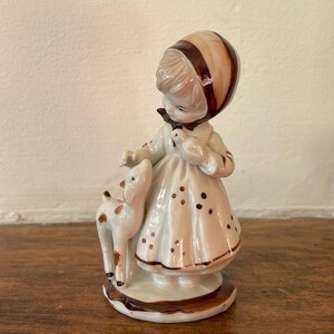 Op de afbeelding: Een porseleinen beeldje van een meisje in een bruin-witte jurk en hoed dat een klein wit kalfje vasthoudt. Het beeldje staat op een bruine voet.