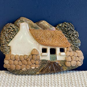 Puede incluir: Figura de cerámica de una cabaña con tejado de paja, paredes blancas y puerta verde. La cabaña está rodeada de muros de piedra y árboles, con un fondo de montaña. La figura se encuentra sobre un fondo azul oscuro.