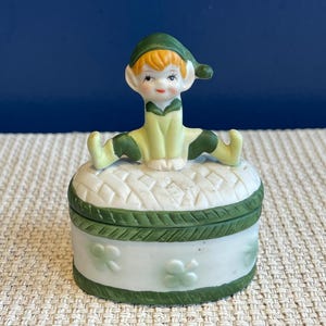 May include: A ceramic trinket box in green and white. The lid has a woven pattern, and the sides are decorated with shamrocks. A small elf figurine, wearing a green hat and outfit, is perched on top of the box.