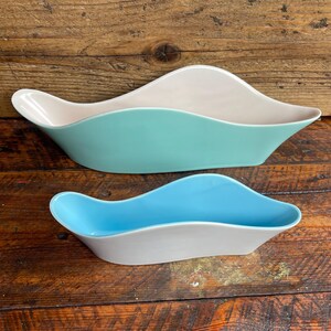 Può includere: Due ciotole in ceramica allungate a forma di onda. La ciotola più grande presenta un esterno liscio color verde menta opaco e un interno color crema. La ciotola più piccola ha un interno blu cielo brillante con un esterno bianco, bordi curvi.
