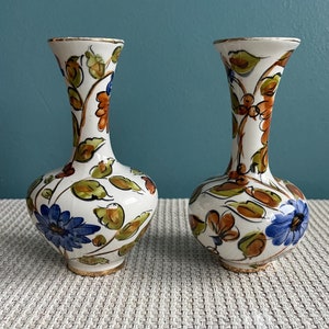 May include: Two small white ceramic vases with a floral pattern in shades of blue, orange, and green. The vases have a gold trim around the top and bottom.