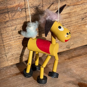 Puede incluir: Títere de burro de madera. El títere tiene un cuerpo amarillo, una sección media roja y pezuñas negras. Tiene una cabeza rosa y marrón con orejas negras y una cola azul. Suspendido por cuerdas.