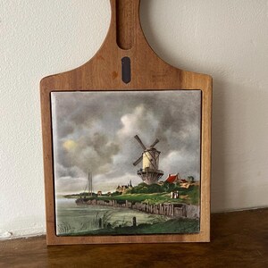 May include: A wooden chopping board with a ceramic tile inset featuring a painting of a windmill in a Dutch landscape. The tile is surrounded by a wooden frame and the board has a handle.