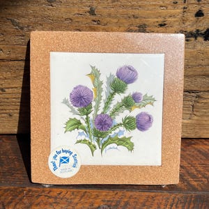 May include: A square coaster with a cork border and a central ceramic tile featuring a botanical illustration of purple thistle flowers with green leaves. A circular sticker in the corner displays the text Thank you for buying Scottish.