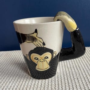 Puede incluir: Una taza de cerámica blanca con un diseño de chimpancé. La taza presenta una cara de chimpancé negra y un asa en forma de brazo. Una mano está pintada en el lateral. La taza está sobre una superficie texturizada.
