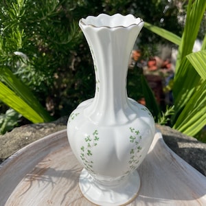 May include: A white porcelain vase with a scalloped rim and a gold-trimmed edge. The vase features a vertical ribbed body decorated with small, delicate green shamrock patterns trailing down the sides from the neck to the base.