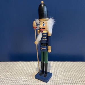 May include: A wooden nutcracker soldier figurine. The figurine has a black hat, blue jacket, green trousers, and black boots. It holds a wooden staff with a red and gold tip. The nutcracker has a white beard and moustache and is standing on a blue base.