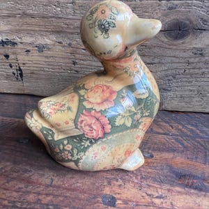 May include: A ceramic duck figurine with a floral pattern in shades of pink, green, and yellow. The duck has a cream-colored beak and head, and the body is adorned with a patchwork design featuring roses and other flowers. The figurine is sitting on a wooden surface.