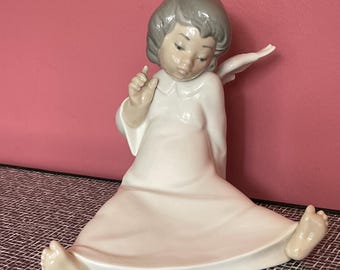 May include: A porcelain figurine of a seated angel with gray hair and a white dress. The angel has a wing on its back and is looking down.