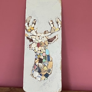 May include: A white wooden board with a mosaic deer silhouette made of colorful ceramic tiles. The deer's antlers are made of white ceramic tiles. The word "STAN" is visible on the deer's body.