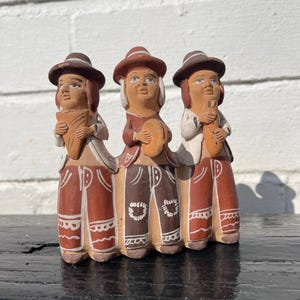 Puede incluir: Figura de cerámica que representa a tres músicos. Cada figura lleva un sombrero marrón, una camisa blanca y pantalones marrones con detalles blancos. La figura central sostiene una pandereta, mientras que las otras sostienen instrumentos de viento. Las figuras están pintadas en tonos tierra.
