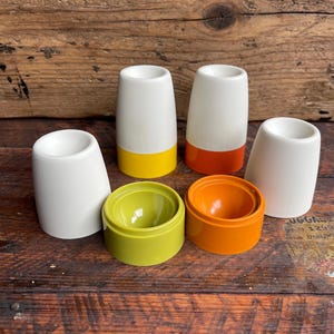 Może przedstawiać: Zestaw sześciu ceramicznych kubków na jajka i uchwytów. Kubki na jajka są białe z kolorowymi podstawami w kolorze żółtym, pomarańczowym i zielonym. Uchwyty są również w kolorze żółtym, pomarańczowym i zielonym. Przedmioty są ułożone na drewnianej powierzchni.