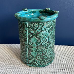 Puede incluir: Un jarrón de cerámica de color turquesa con un patrón floral en relieve decorativo. El jarrón tiene una parte superior redondeada con un borde ligeramente acampanado.