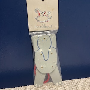 May include: A wooden elephant toy in a clear plastic bag. The elephant is light grey with blue accents and gold-coloured rivets. The packaging includes a logo of a rocking horse and the text "THE TOY WORKSHOP".