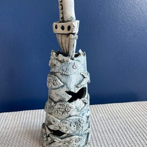May include: A tall, textured ceramic sculpture in shades of white and blue. The cylindrical form features carved fish and leaf designs, with a central cutout. The top has a crown-like structure and a vertical element.