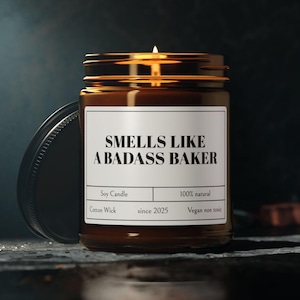 May include: A lit soy candle in a brown glass jar with a black lid. The label reads "SMELLS LIKE A BADASS BAKER." Additional text includes "Soy Candle," "Cotton Wick," "100% natural," "since 2025," and "Vegan non toxic."