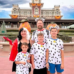 May include: A family dressed in costumes featuring a person in a black dress with a red coat and black and white wig, and three people in white t-shirts with black spots, red collars, and yellow tags, wearing matching dalmatian ear headbands.
