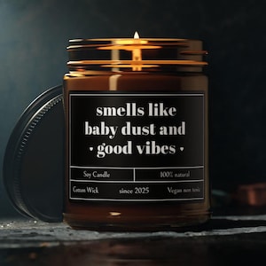 Puede incluir: Una vela de soja encendida en un frasco de vidrio marrón con una etiqueta negra que dice "smells like baby dust and good vibes". La vela tiene una mecha de algodón y está etiquetada como vegana y no tóxica.