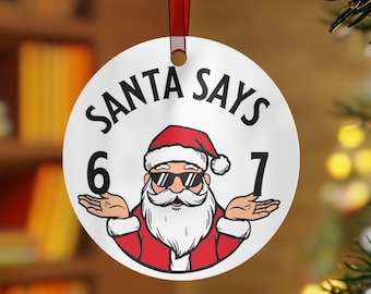 May include: A round Christmas ornament with a red ribbon. The ornament features a cartoon Santa Claus wearing sunglasses and the text "SANTA SAYS" above the image. The numbers 6 and 7 are on either side of Santa.