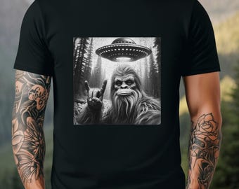May include: Black and white graphic t-shirt featuring a Bigfoot with a rock and roll hand gesture, with a UFO hovering above him in a forest setting.