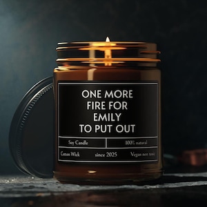 Puede incluir: Una vela de soja en un tarro de vidrio marrón con tapa dorada. La etiqueta dice "ONE MORE FIRE FOR EMILY TO PUT OUT." Texto adicional: "Soy Candle", "100% natural", "Cotton Wick", "since 2025" y "Vegan non toxic."